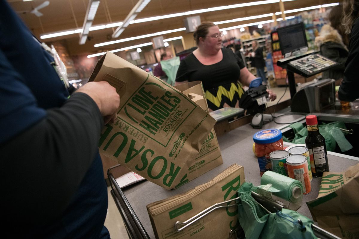 At the end of a black conveyor belt with the interior of a grocery store in the background, a person in a blue sweater holds a brown paper bag with green writing.
