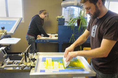 Jessica Hettich, left, lays shirts on the dryer while Blue Button Apparel  director Scott Ellis screens designs on them at the  Hillyard shop recently.  Ellis hopes that more young people like Hettich, a Lewis and  Clark sophomore, can work at, and be mentored by, his company.  (Jesse Tinsley / The Spokesman-Review)