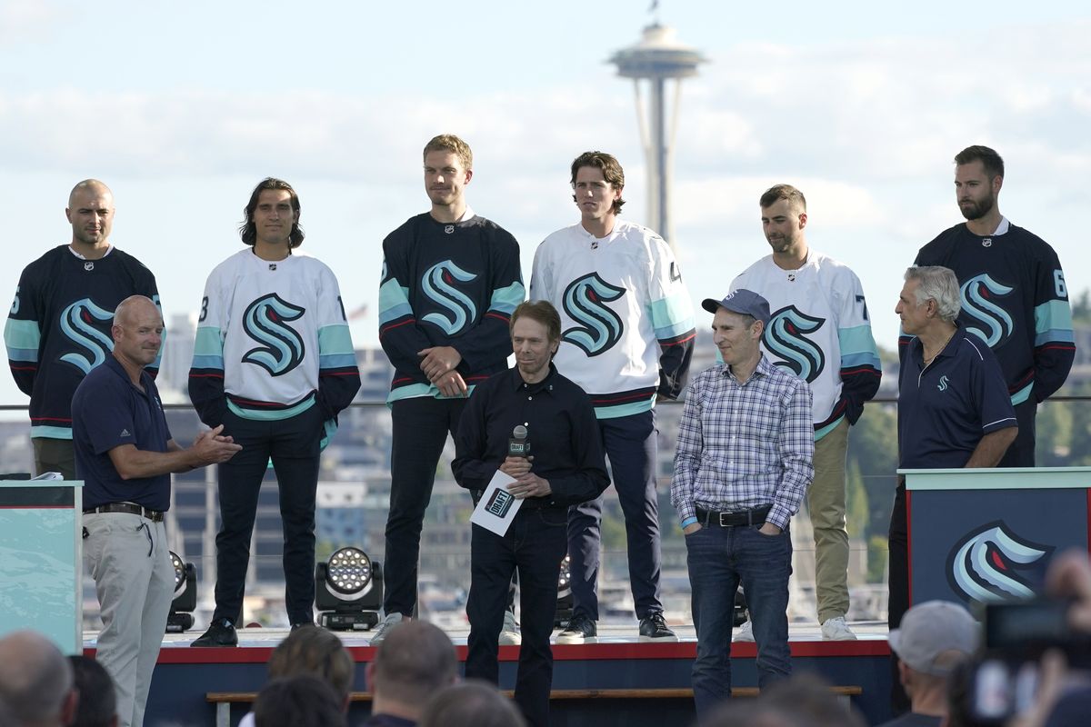 New Seattle Kraken NHL hockey players, back row from left, Mark Giordano, Brandon Tanev, Jamie Oleksiak, Hadyn Fluery, Jordan Eberle and Chris Dreidger stand on stage with Kraken owners David Wright, front left, Jerry Bruckheimer, front center, and Andy Jassy, front second from right, and Kraken general manager Ron Francis, front right, Wednesday, July 21, 2021, after being introduced during the Kraken