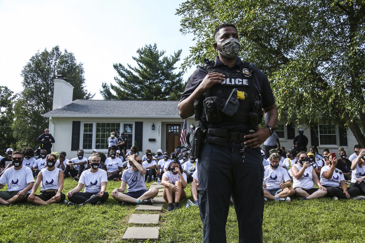 In this image from video, a Louisville Metro Police Department office stands guard outside the home of Kentucky Attorney General Daniel Cameron as protestors sit in his front yard in Louisville, Kentucky Tuesday, July 14, 2020. About two dozen protestors were arrested. Protesters were chanting Breonna Taylor