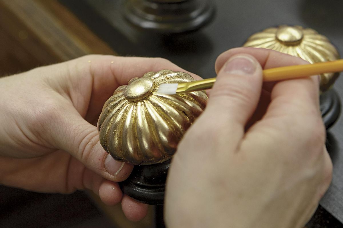 In this photo provided by Ethan Allen, a craftsman uses a small artists brush to press the gold leafing material into the detailed carved elements of the finial on an Ethan Allen Georgetown Bed in their North American workshops. (Ethan Allen)