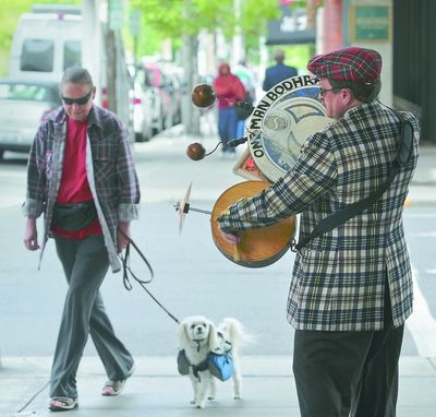 Steven Schneider, 54, performs his one-man-band act as Jo Smittle, 62, and her dog, Zoe, pass him on the corner of Howard Street and Main Avenue during the first day of Spokane Street Music Week in downtown Spokane on Monday.  Schneider says if you want people to give money, “You have to look ’em right in the eye.” (Dan Pelle)