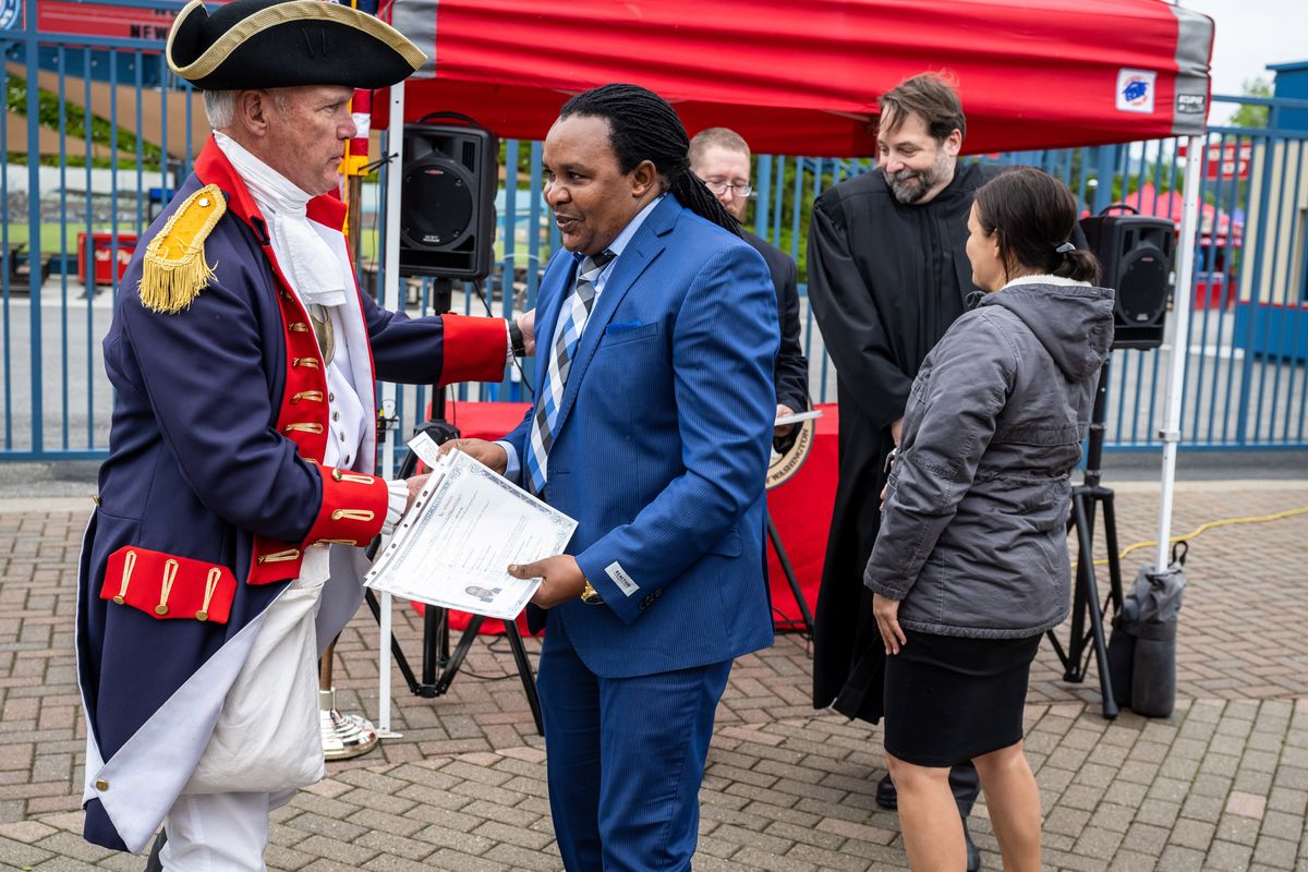 After receiving his Certificate of Naturalization from U.S. Magistrate Judge James Goeke (in back), Stephen Gichure is handed an American flag pin from Stan Wills with the Sons of the American Revolution, during a U.S. Citizenship and Immigration naturalization ceremony Tuesdya held at Avista Stadium.  (COLIN MULVANY/THE SPOKESMAN-REVI)