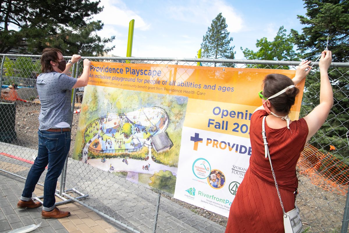 Josh Morrisey, marketing assistant (left) and Fiona Dickson, communications manager for Spokane Parks and Recreation, hang the visual rendering sign for the Providence Playscape to the chainlink fence surrounding its perimeter in Riverfront Park on June 24, 2020. The playground is geared towards children of all needs in an effort to boost equity and inclusivity and is slated to open in fall 2020.   (Libby Kamrowski/ THE SPOKESMAN-REVIEW)