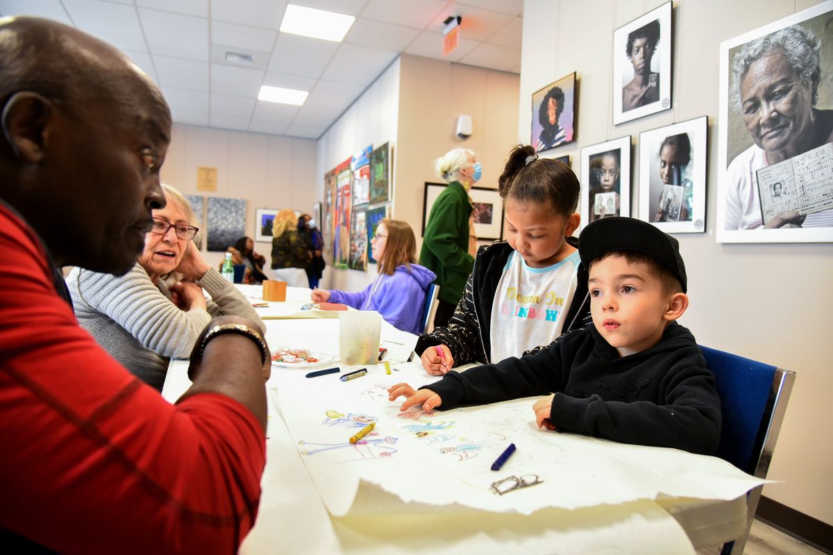 Surrounded by photos, paintings and prints that are part of the "Our Stories Our Voices" exhibit, African artist Sironka, left, talks about art with Elijah Bivens, 5, right, Shay Love, 6, second from right, and Kay Bivens, second from left. The exhibit opened Saturday afternoon to young people before a formal opening reception at the Carl Maxey Center.  (Jesse Tinsley/The Spokesman-Review)