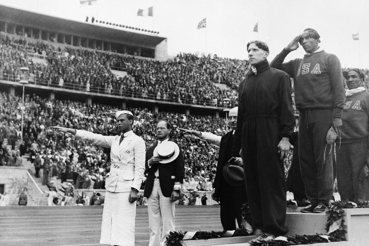 Gold medalist Jessie Owens, second right, salutes during the playing of the national anthem in the medal ceremony of the 100-meter final in Berlin on Aug. 3, 1936. Silver medalist Tinus Osendarp, third from right, of Holland, and bronze medalist Ralph Metcalf, right, listen, along with a matron who holds a Nazi salute. The 1936 Games in Berlin, awarded about two years before Adolf Hitler became dictator, went ahead under Nazism. The documentary, “The 1936 Olympic Games: Nazi Secrets at the Berlin Olympics” looks at the negotiations to bring the games to Berlin.  (Associated Press)