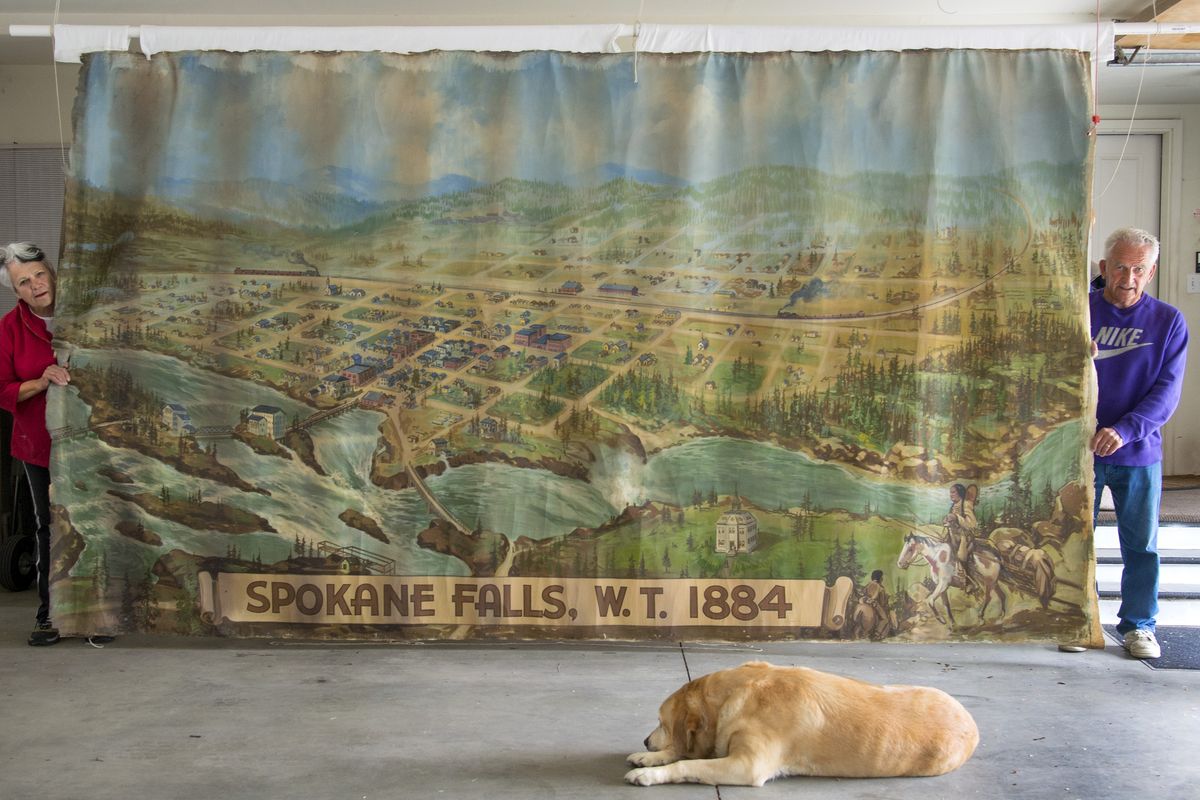 Dinah and Boyd Carlson display an oil-on-canvas mural that may have once decorated a Spokane bank. Tank the dog sits in the foreground. The mural is an artist’s rendition of the 1884 Bird’s Eye View of Spokane Falls, and was painted in 1938. It measures 149 inches by 89 inches. (Colin Mulvany)
