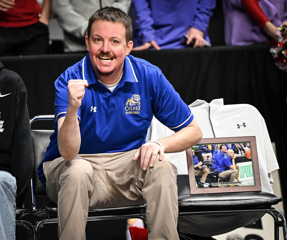 New Colfax boys basketball coach Ben Aune cheers a basket by his Bulldogs team as he sits next to a framed photo of former head Colfax boys basketball coach Reece Jenkin who died last Friday from pancreatic cancer. Colfax won its game on Thursday at the Arena.  (Colin Mulvany/The Spokesman-Review)