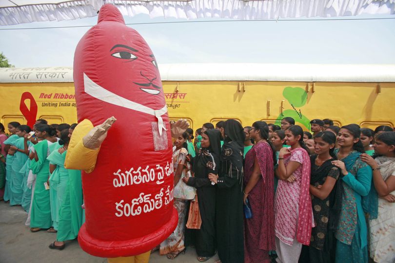 A person dressed as a giant condom walks past visitors standing in a queue to visit the Red Ribbon express train, background, at a railway station in Hyderabad, India, Thursday, April 15, 2010. The Red Ribbon express train is traveling across the country as part of a campaign to spread awareness on HIV and AIDS. (Mahesh A / Associated Press)