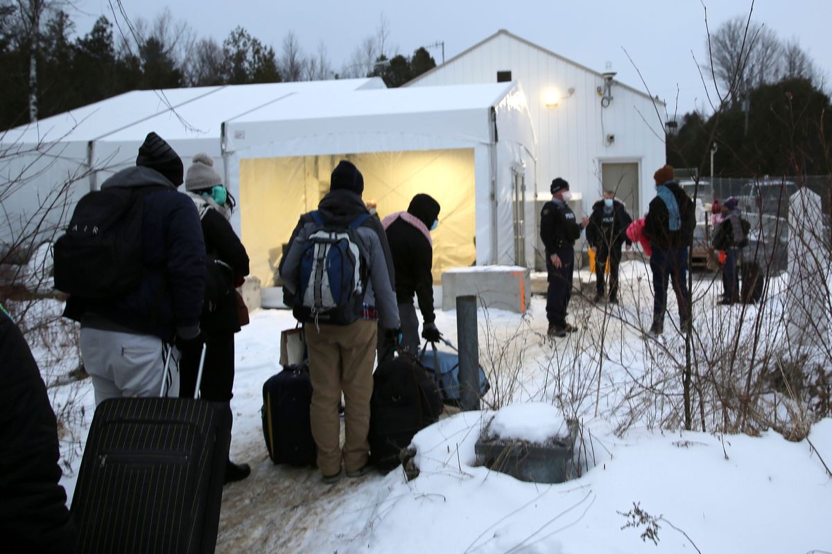 Migrants line up on the border of the United States, foreground, and Canada, background, at a reception center for irregular borders crossers, in Saint-Bernard-de-Lacolle, Quebec, Canada, Wednesday Jan. 12, 2022, in a photo taken from Champlain, N.Y. They are crossing the U.S.-Canadian border into Saint-Bernard-de-Lacolle, Quebec, where they are arrested by the Royal Canadian Mounted Police and then allowed to make asylum claims. The process was halted for most cases after the 2020 outbreak of COVID-19, but the Canadian government changed its policy in November, allowing the process to continue.  (Wilson Ring)