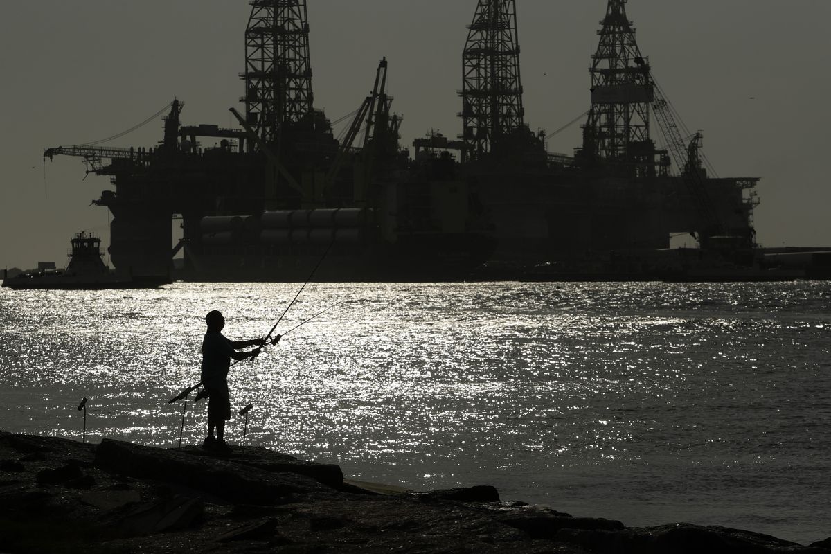 FILE - A man wears a face mark as he fishes near docked oil drilling platforms, Friday, May 8, 2020, in Port Aransas, Texas. The U.S. Interior Department on Wednesday, Nov. 17, 2021, is auctioning vast oil reserves in the Gulf of Mexico estimated to hold up to 1.1 billion barrels of crude. It