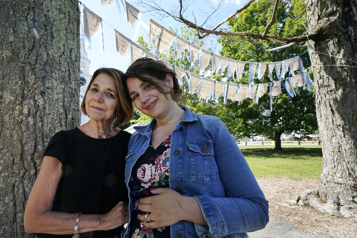 Widow Marcy Jacobs, left, stands with her daughter, Jaclyn Winer, under flags bearing names of people, including her husband, Keith Jacobs, who have died from COVID-19, outside the First Congressional Church, Thursday, June 17, 2021, in Holliston, Mass. The flags are part of the COVID Art and Remembrance project spearheaded by Jaclyn. “Don’t expect us to move on without giving us a place to grieve,” Marcy Jacobs said, recalling her husband as kind, uncomplaining and simple. “Is it a stone for everyone? I don’t know."  (Elise Amendola)