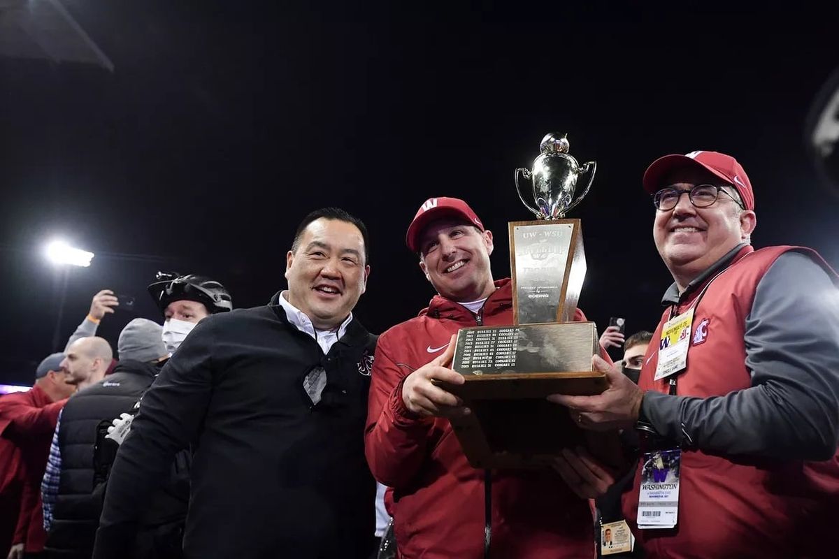 Washington State football coach Jake Dickert hoists the Apple Cup given to him by Washington Gov. Jay Inslee (not pictured), as WSU athletics director Pat Chun, left, and President Kirk Schulz smile after WSU defeated the Washington Huskies 40-13 on Nov. 26, 2021, at Husky Stadium in Seattle.  (Tyler Tjomsland/Spokane Daily Chronicle)