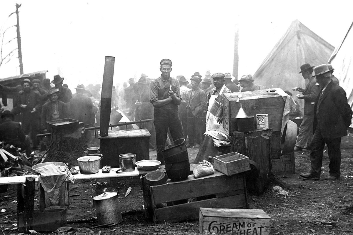 This is a 1910 scene of firefighters cooking breakfast at camp. The man in the apron is Pat Grogan, leader of the men who died on Storm Creek. A hazy outline of the Avery Ranger Station is in the background.   (Courtesy Idaho / Photo courtesy of the Museum of North Idaho)