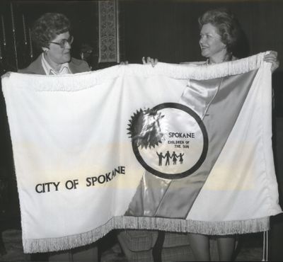 Mrs. W. Tom Mableson, left, and Mrs. Terry H. Tate display the 1977 official city flag that was created by members of the Spokane Falls Needlework Guild. The white velvet background is colored by a chartreuse and an aqua strip and bears the city logo. The flag was designed by Lloyd Carlson, who also created the Expo ’74 mobius strip symbol.  (Spokesman-Review archives)