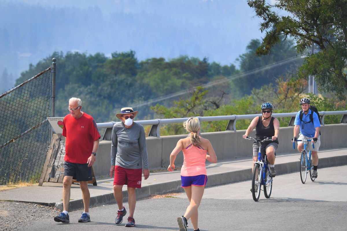 Dan Schnell and Bill McElroy, wearing the mask, cross paths with a runner and a pair of cyclists on the Centennial Trail during a smoky noon hour Thursday, Aug. 16, 2018, in Spokane. “We walk everyday that starts with “T,” said Schnell. (Dan Pelle / The Spokesman-Review)