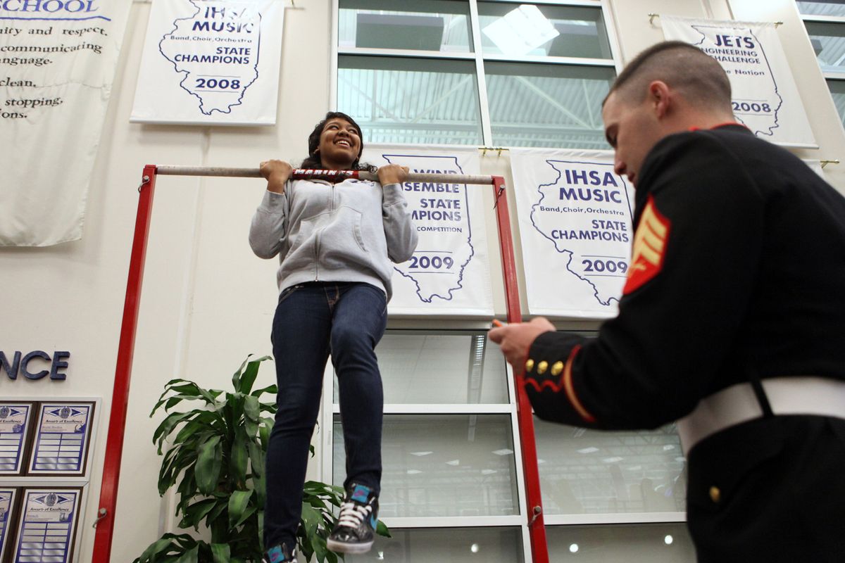 Marine Corps Sgt. David Pope, right, keeps track of time while senior Angelica Moss, 18, hangs from the pull-up bar outside the lunchroom at Vernon Hills High School in Vernon Hills, Ill., in May. The Marines recruit at Vernon Hills High School by setting up a pull-up bar to lure students over to their table during lunch. The students can win  prizes based on how well they do. 