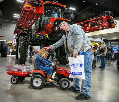 Bill Black, of Moses Lake, pats his granddaughter Fae Brooks, of Spokane, atop the head as she gets behind the wheel of an electric toy Case tractor next to a full-size Case 4440 Patriot sprayer during their visit to the Spokane Ag Show on Feb. 2, 2022, in the Spokane Convention Center. Black said he is not a farmer but is “a farmer at heart.”  (DAN PELLE/THE SPOKESMAN-REVIEW)