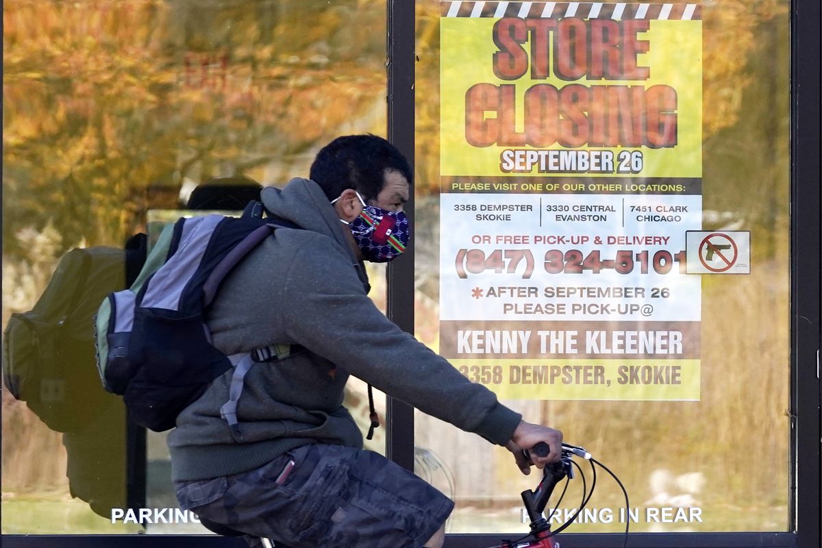 FILE - In this Friday, Nov. 6, 2020, file photo, a man wearing a face mask rides a bicycle past a closed store in Wilmette, Ill. States in the U.S. are renewing their push for more federal money to deal with the fallout from the coronavirus outbreak and to help them distribute a vaccine when one becomes widely available sometime in 2021.  (Nam Y. Huh)