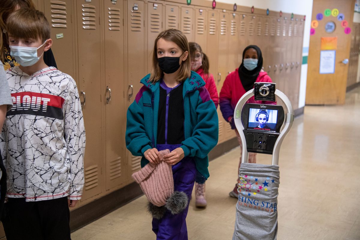 Fourth-grader Juliet McLemore, center (holding her hat) performs the duties of "bot buddy" and looks out for obstaces while she walks beside classmate Madelyn Schible , right, who appears on the screen of her "robot" while it rolls through the halls at Madison Elementary School Monday, Dec. 6, 2021 in Spokane, Washington. Schible is able to attend school via the robot, which can navigate through hallways and put her in whichever classroom she needs to be in throughout the day. Madelyn, who is disabled by cerebral palsy, can drive the robot from her laptop at home. Because of her fragile health, she attends school with the robot, which was provided by Wishing Star, a charity that helps kids with terminal or very complex illnesses. Wishing Star only has a handful of the machines and only provides them to children who have medically qualified through their screening process. Above Madelyn's screen is a camera, which she can tilt up or down while driving through the halls.  (Jesse Tinsley/THE SPOKESMAN-REVI)