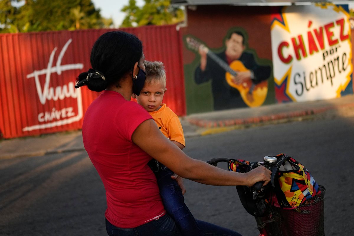 A woman rides a bike with her son past a mural of the late Venezuelan President Hugo Chavez in his hometown Sabaneta, in Barinas state in Venezuela, Friday, Jan. 7, 2022. The country