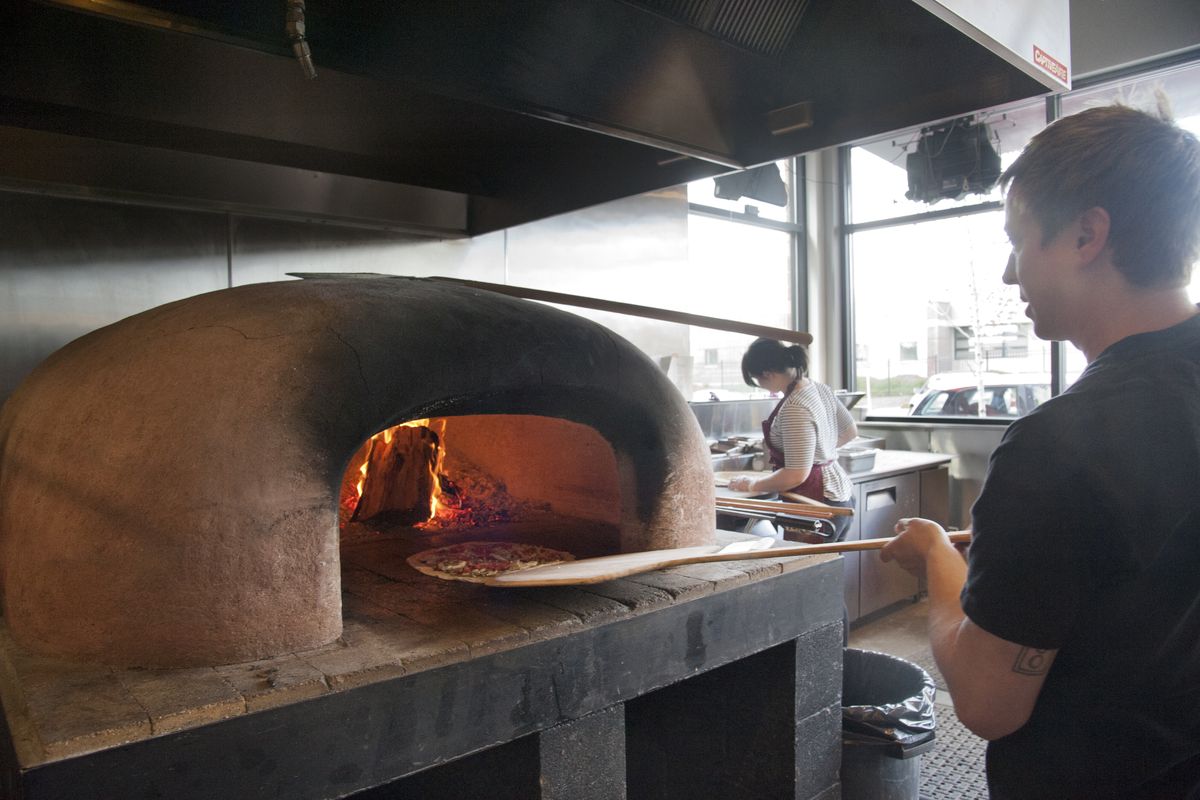 Seth Carey slides a pizza into the wood-fired clay oven Tuesday at Veraci Pizza in Kendall Yards. Veraci Pizza began as a mobile cart at area farmers markets before opening its restaurant in 2014. A farmers market will begin next month outside the restaurant. (Jesse Tinsley)