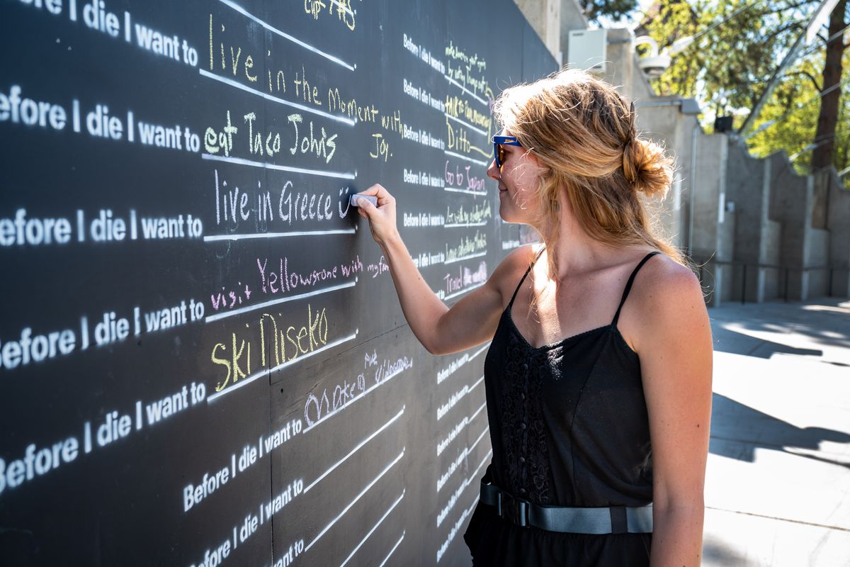 Hansine Holm writes that she wants to live in Greece on the “Before I Die” wall at the U.S. Pavilion in Riverfront Park on Aug. 4. Passersby can pick up a piece of chalk and fill in one of 96 blanks with something they want to do during their respective lifetimes.  (Colin Mulvany/THE SPOKESMAN-REVIEW)