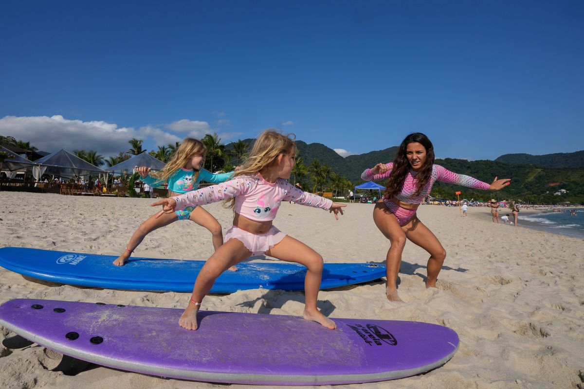 Rayana Tanimoto teaches her five-year-old twin daughters Eloa, left, and Ayla, how to surf, at Maresias beach, in Sao Sebastiao, Brazil, Saturday, Nov. 27, 2021. Tanimoto, caught the surfing bug from her father. Together, they opened a small hotel on Maresias beach so she could raise her daughters, make a living and get in the water as often as possible.  (Andre Penner)