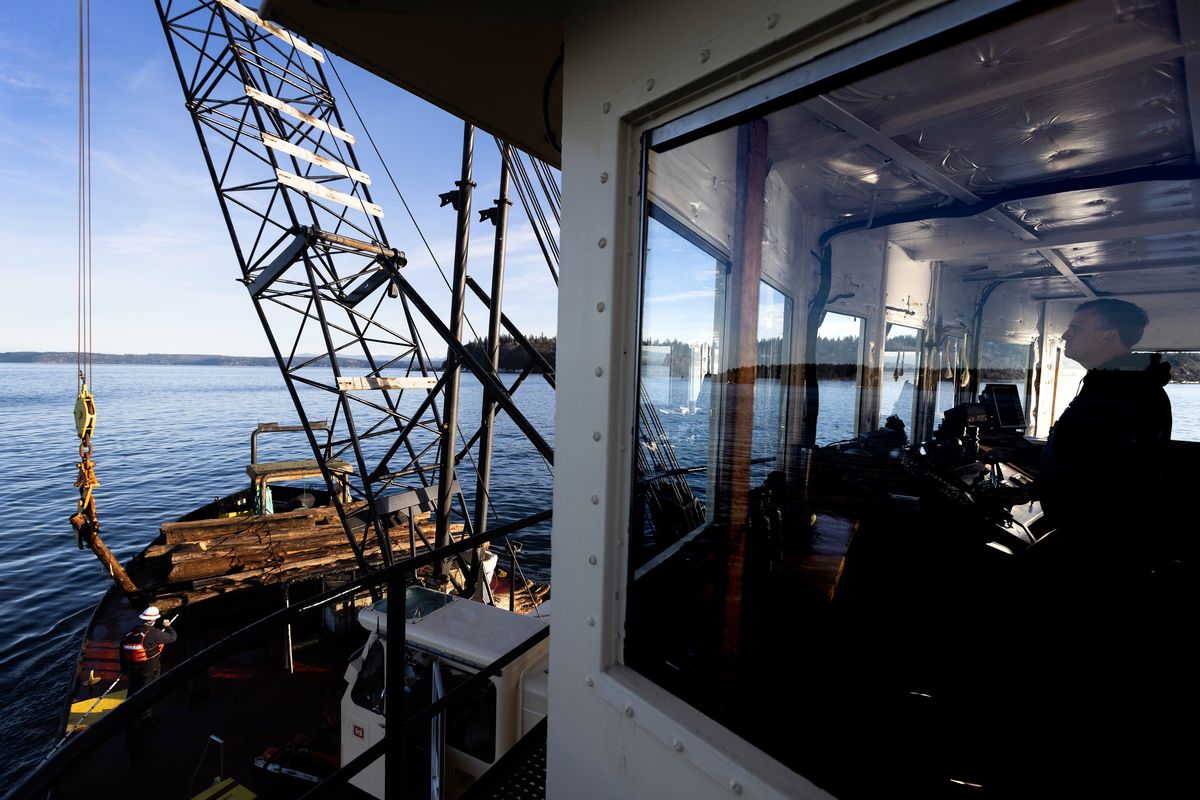 Skip Green, right, captain of the M/V Puget, maneuvers the boat while a crane operator below works to bring aboard a log, Tuesday, Jan. 10, 2023, as the U.S. Army Corps of Engineers debris recovery vessel goes out on Puget Sound to pick up stray floating logs and things that could be a hazard to boat traffic.    (Ken Lambert/The Seattle Times/TNS)