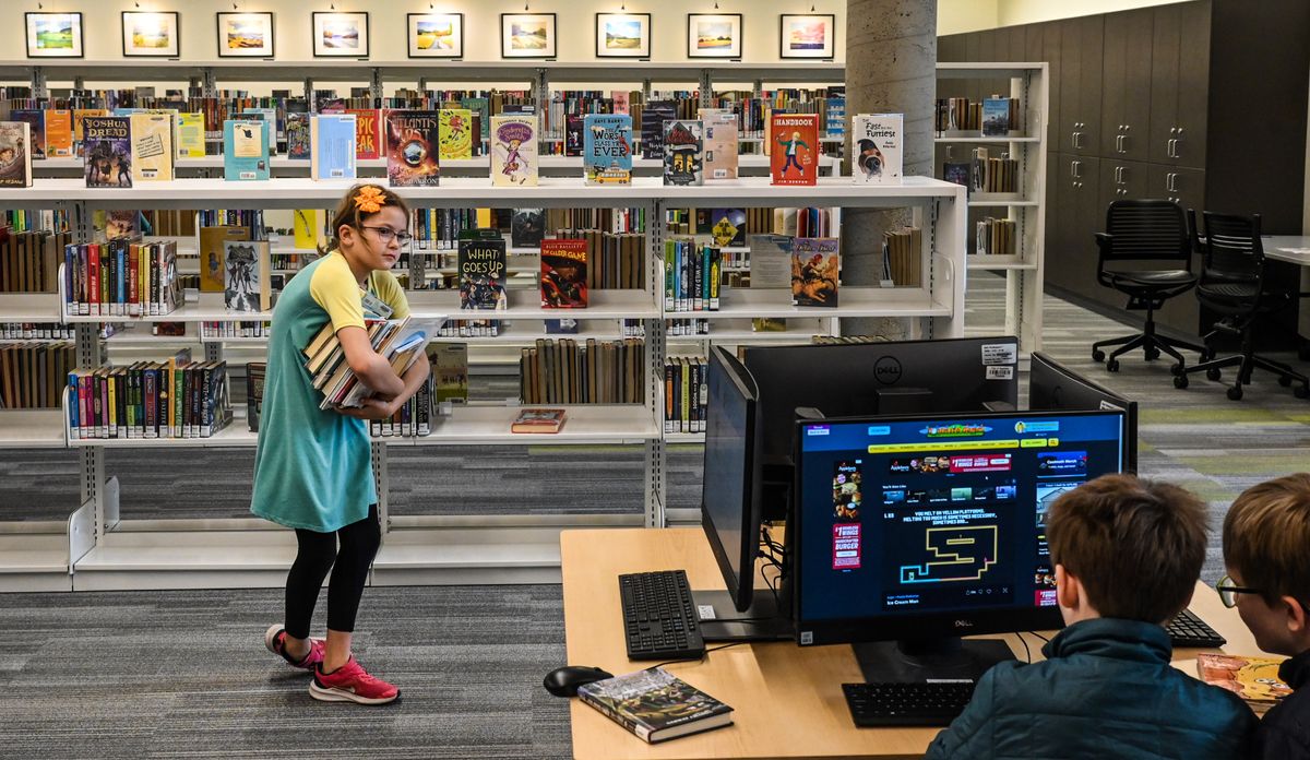 Eleanor Culp, 9, has an armful of books she intends to read as brothers James, 8, and Archie Donahoe, 10, play a game in the new children’s area of the South Hill Library on April 6. Along the back wall is an area for local artists to display their work. Last month’s opening of the South Hill and Indian Trail libraries marks the end of major construction resulting from a $77 million bond approved by voters in 2018.  (DAN PELLE/THE SPOKESMAN-REVIEW)