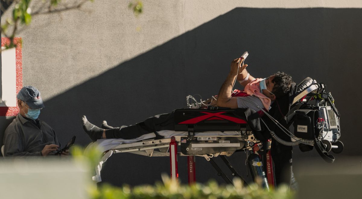 An unidentified patient uses his mobile phone while receiving oxygen on a stretcher, as Los Angeles Fire Department Paramedics monitor him outside the Emergency entrance, waiting for his room at the CHA Hollywood Presbyterian Medical Center in Los Angeles Friday, Dec. 18, 2020. Increasingly desperate California hospitals are being “crushed” by soaring coronavirus infections, with one Los Angeles emergency doctor predicting that rationing of care is imminent.  (Damian Dovarganes))