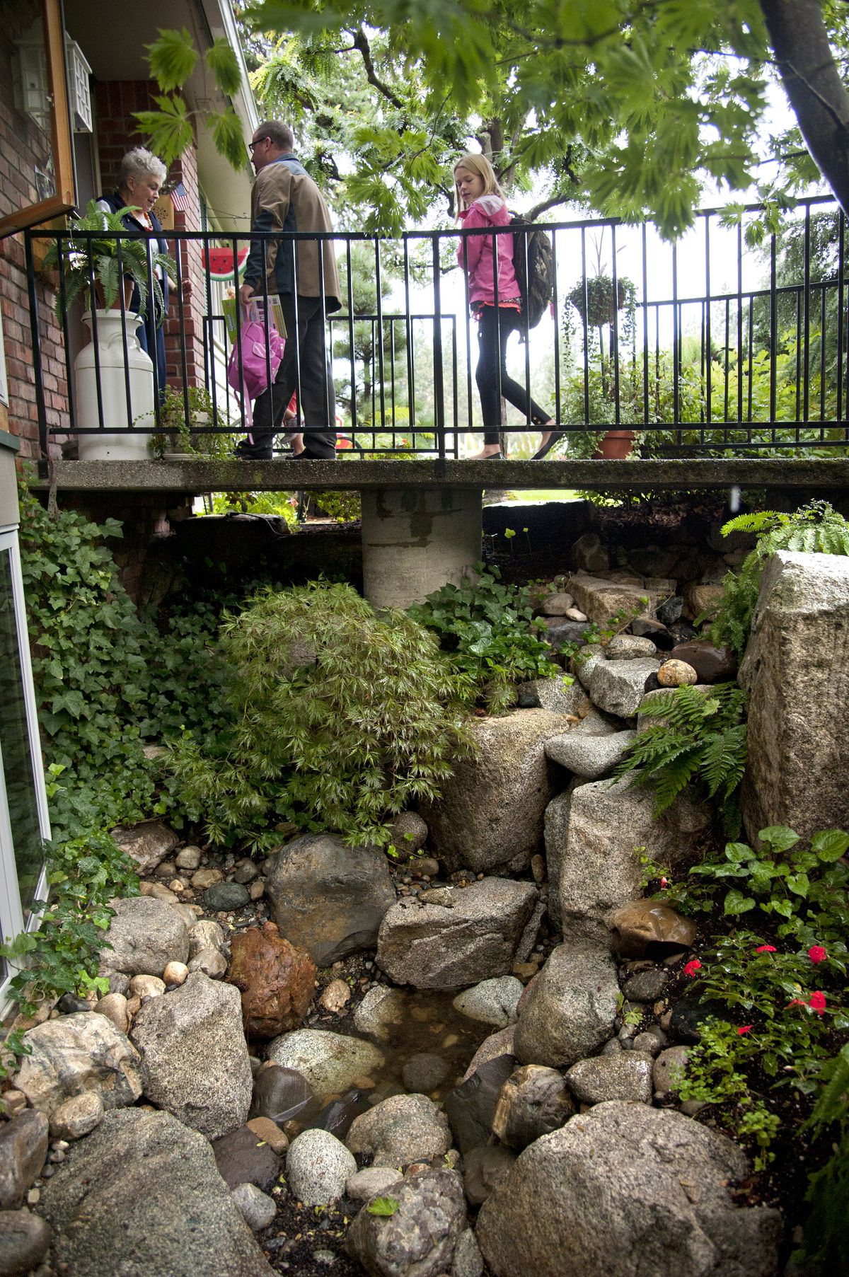 Day care provider Liz Kocherhans greets Jason Harris and his daughters Kendall Harris, 4, hidden, and Kaitlyn Payne, 10, right, as they cross over the water feature in front of her northwest Spokane home on Thursday. Kocherhans was ordered by the Department of Early Learning to shut the water feature off and drain it during business hours. (Dan Pelle)