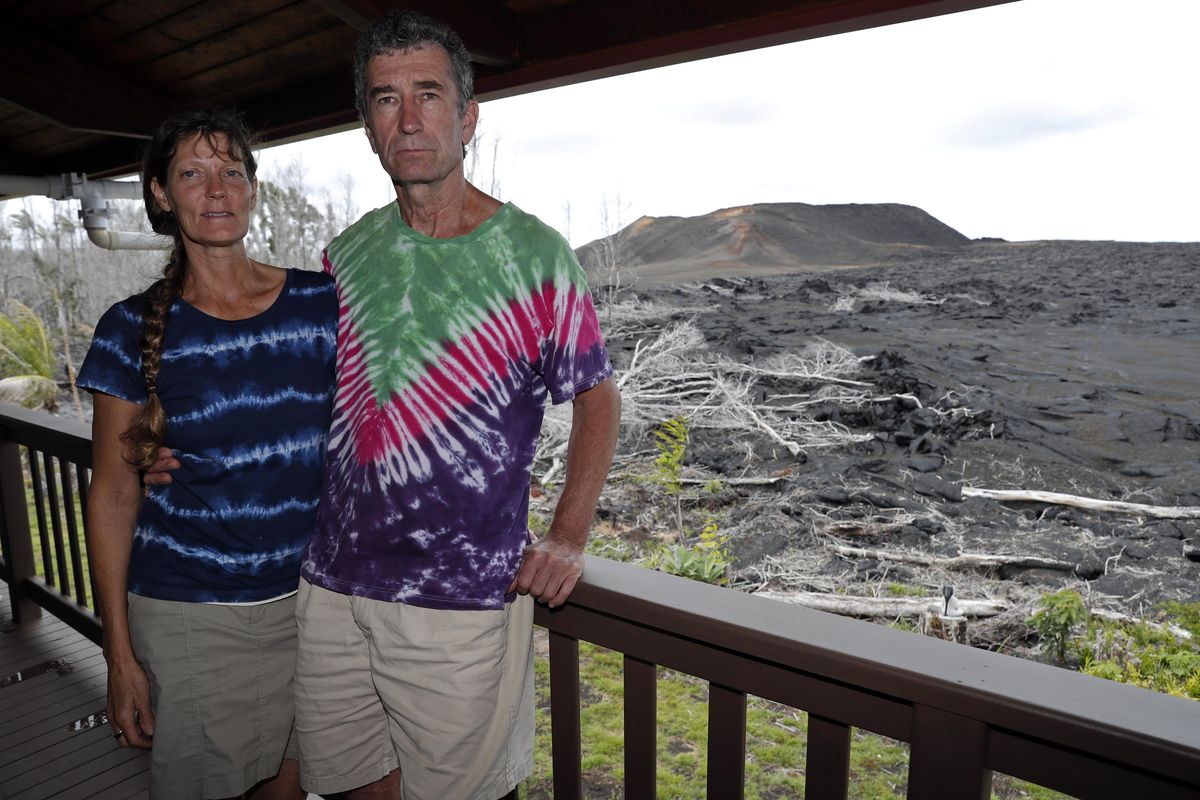 On Tuesday, April 23, 2019, with a now-dormant towering cinder cone looming in the background, Mark and Jennifer Bishop stand on the deck of their home near Pahoa, Hawaii, which was spared by the lava. The epicenter of the 2018 eruption – one of more than 20 places where the ground split open and released massive explosions of molten rock – is now in their front yard. The red-hot fluid oozed onto their property and stopped about 20 feet from the home. Theirs is now the last house on the street. (Marco Garcia / Associated Press)