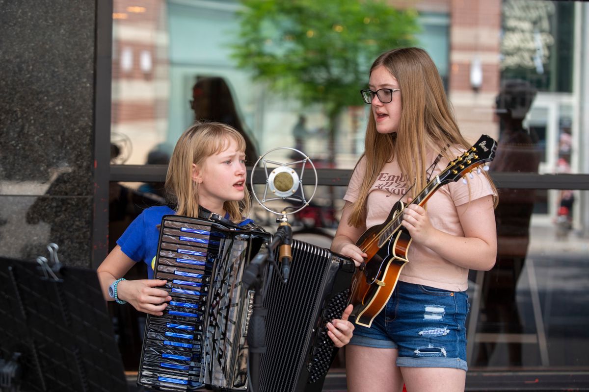 Sisters Ivy Eyer, 12, left, and Neilia Eyer, 14, play and sing harmony at Street Music Week – the annual rally of musicians who spread out through the lunch hour to raise money for Second Harvest food bank – shown Monday on Main Avenue in downtown Spokane. The two have been performing at the annual gathering for almost 10 years, and their father, Cary Eyer, has stepped in to help organize the event after the passing of organizer Jim Lyons.  (Jesse Tinsley/THE SPOKESMAN-REVIEW)