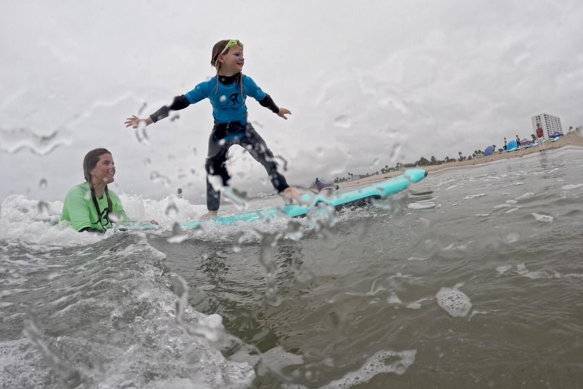 With a push and balance assistance from instructor Genevieve Grossmueller, surfer Sienna Cohee rides a wave together at Aqua Surf surf School at Santa Monica State Beach in Santa Monica Tuesday, June 24, 2025. Aqua Surf School