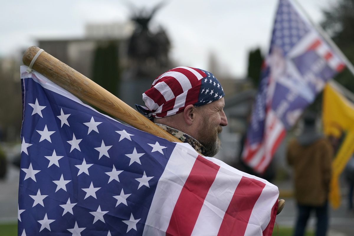 A man carries a U.S. flag attached to a baseball bat as he waits Wednesday at the Capitol in Olympia, before the start of a protest rally against the counting of electoral votes in Washington, D.C., affirming President-elect Joe Biden’s victory.  (Ted S. Warren)