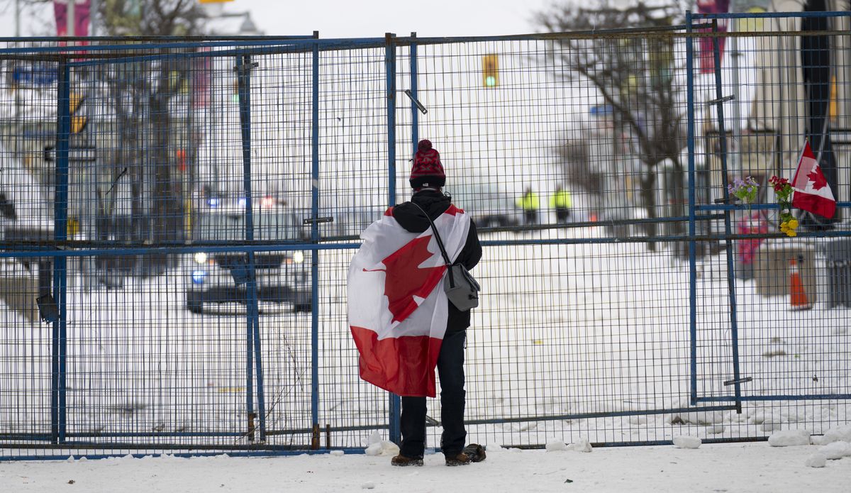 A lone protester stands draped in the Canadian flag at a temporary fence controlling access to streets near Parliament, in Ottawa, Sunday, Feb. 20, 2022.  (Adrian Wyld)