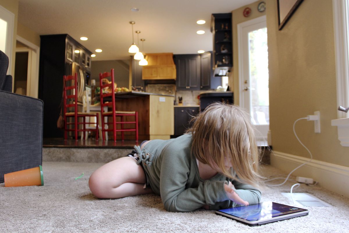 Lizzie Dale sprawls on the floor to play games on an iPad as her siblings work on school work in the kitchen behind her in their home in Lake Oswego, Ore., Oct. 30, 2020. In Oregon, one of only a handful of states that has required a partial or statewide closure of schools in the midst of the COVID-19 pandemic, parents in favor of their children returning to in-person learning have voiced their concerns and grievances using social media, petitions, letters to state officials, emotional testimonies at virtual school board meetings and on the steps of the state