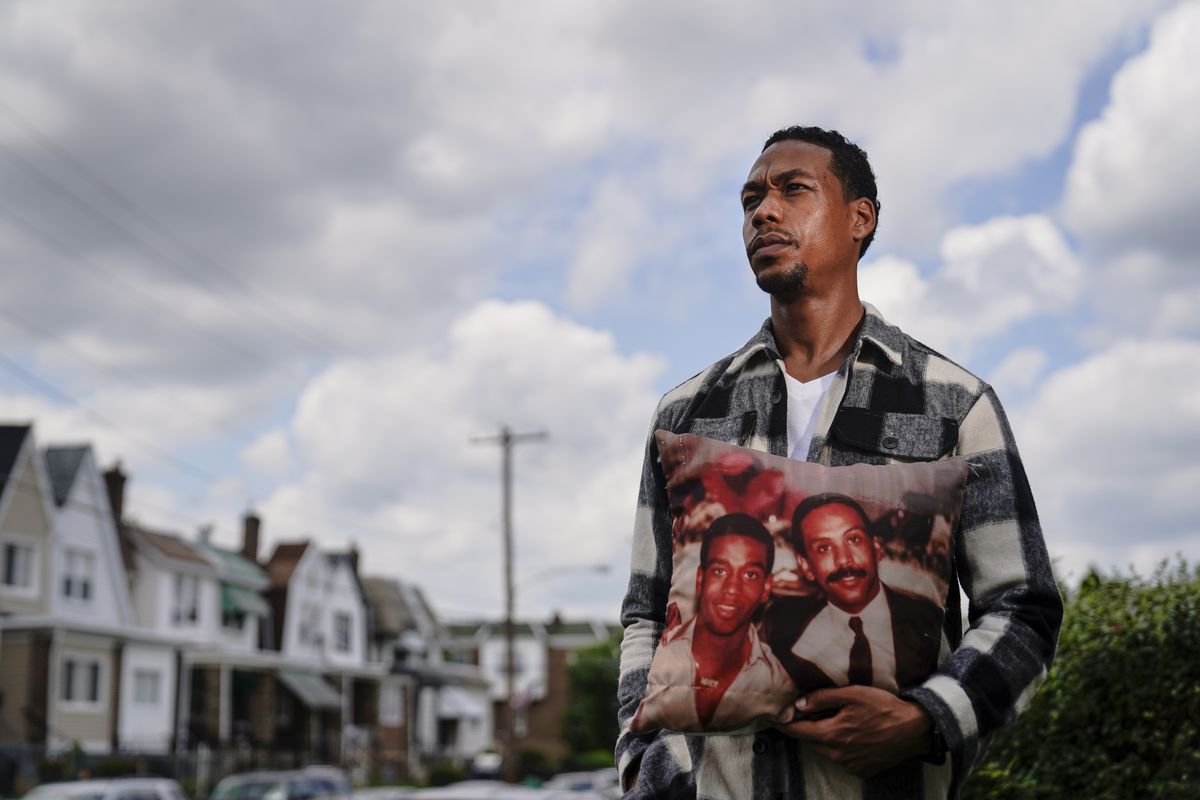 In this July 9, 2021, photo Brett Roman Williams poses for a photograph while holding a pillow with a photo of his father, Donald Williams, lower right, and brother Derrick Williams who both were killed by gunfire 20 years apart, in Philadelphia.  (Matt Rourke)