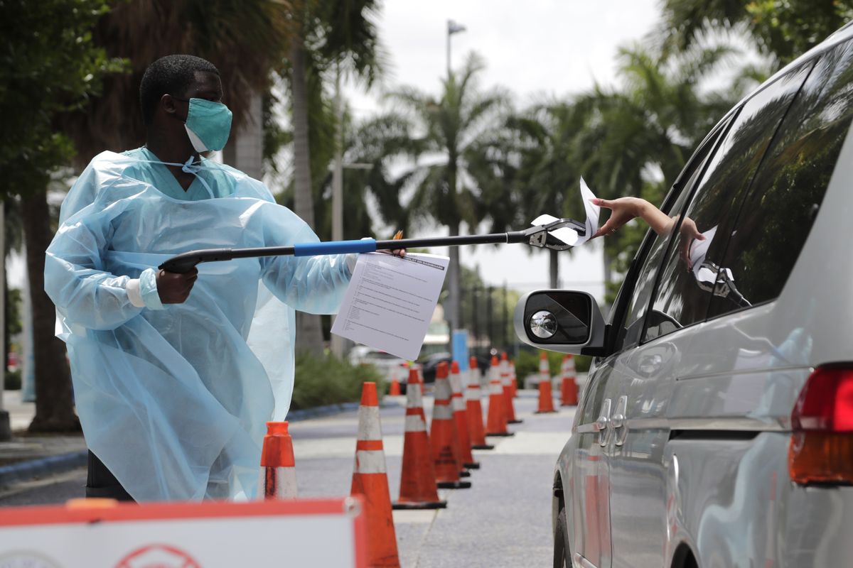 Healthcare worker Dante Hills, left, passes paperwork to a woman in a vehicle at a COVID-19 testing site outside of Marlins Park, Monday, July 27, 2020, in Miami. The Marlins home opener against the Baltimore Orioles on Monday night has been postponed as the Marlins deal with a coronavirus outbreak that stranded them in Philadelphia.  (Lynne Sladky)