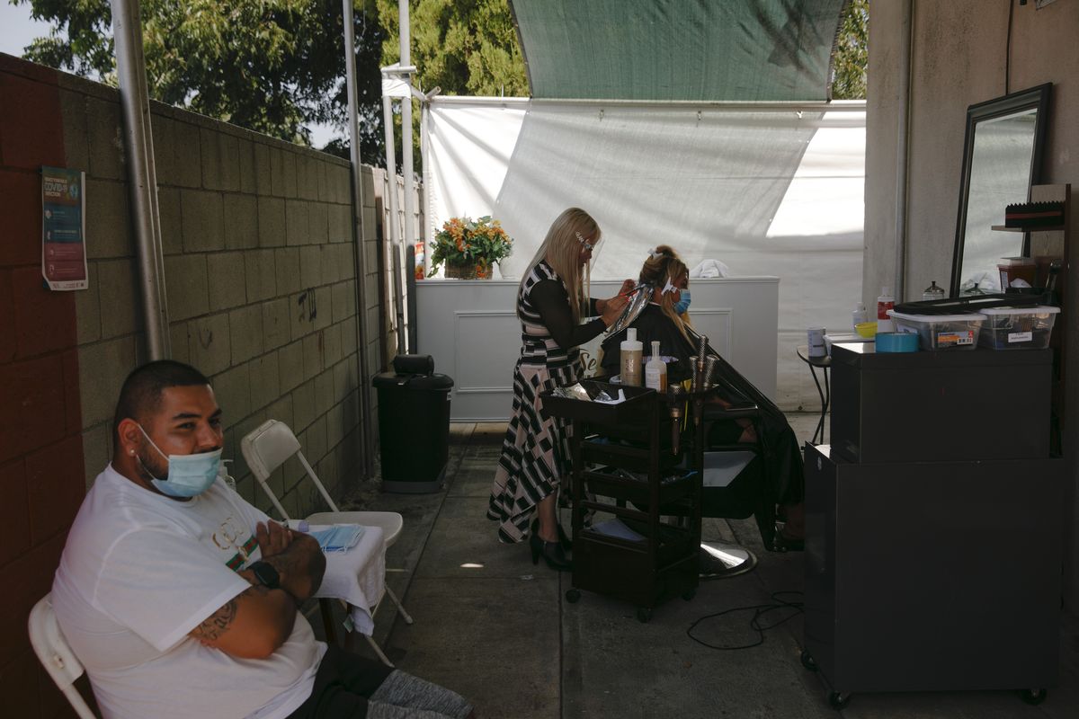 Mario Perez, 32, left, sits on a chair while his wife, Jocelyn, gets her hair bleached at an outdoor hair salon in the Watts neighborhood of Los Angeles, Monday, Aug. 3, 2020. Watts has changed demographically from an exclusively Black neighborhood in the 