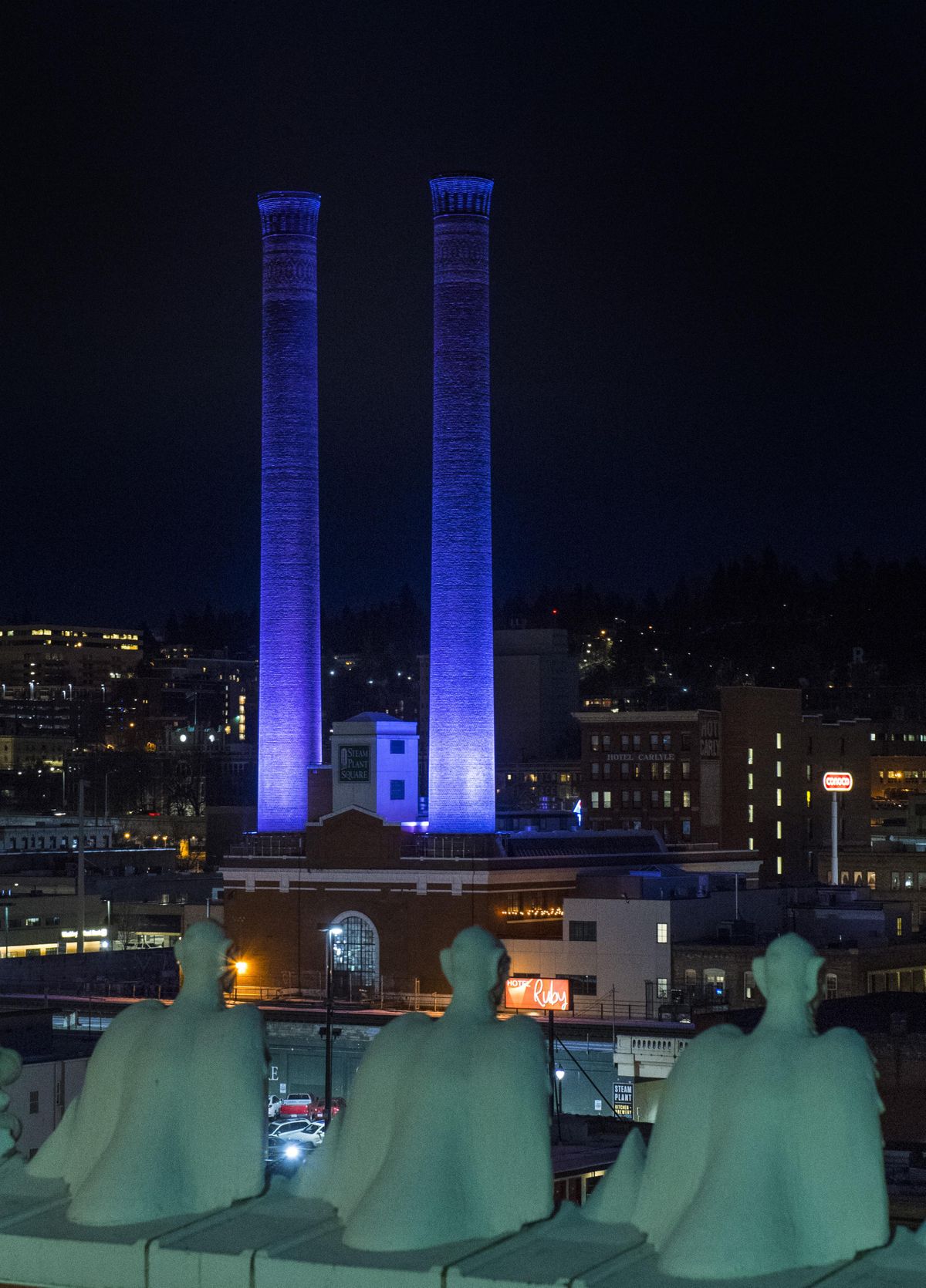 The gargoyles atop the Chronicle building keep watch over the Steam Plant smoke stacks bathed in blue light in February 2018. (Colin Mulvany / The Spokesman-Review)