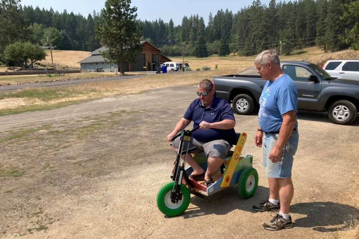 Scott Hendricks test-rides a Rainbow Freedom Cart as Inland Northwest Trinity Project volunteer Ron Bohman looks on in July. Hendrick’s pandemic project is reaching worldwide. He makes wooden crosses that are installed in the rear of the carts built by the Trinity Project and shipped overseas to people in need of personal mobility scooters.  (Courtesy)