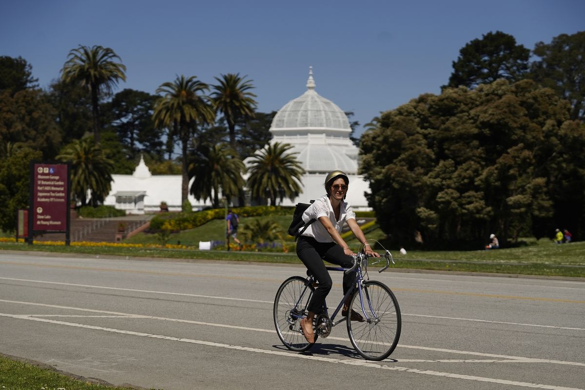A woman on a bicycle rides along the car-free John F. Kennedy Drive in Golden Gate Park with the Conservatory of Flowers in the background, Wednesday, April 28, 2021, in San Francisco. At the start of the pandemic, San Francisco closed off parts of a major beachfront highway and Golden Gate Park to cars so that people had a safe place to run and ride bikes. Open space advocates want to keep those areas car-free as part of a bold reimagining of how U.S. cities look. But opponents decry the continued closures as elitist, unsafe and nonsensical now that the pandemic is over and people need to drive again.  (Eric Risberg)