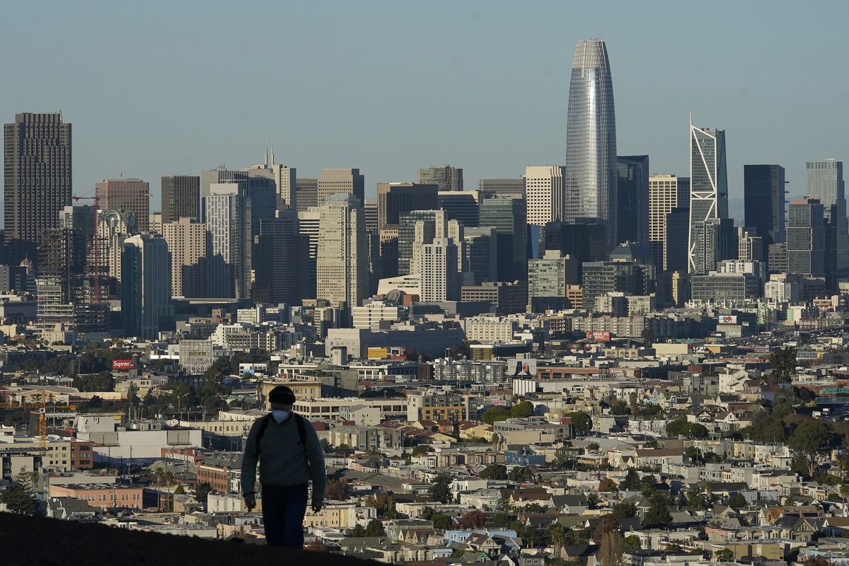 FILE - In this Dec. 7, 2020, file photo, a person wearing a protective mask walks in front of the skyline on Bernal Heights Hill during the coronavirus pandemic in San Francisco. California