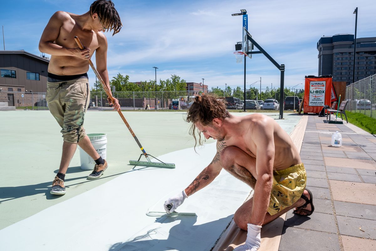 Mural artist Joshua Martel, on right, kneeling, with helper Greyson Hatcher spread a base coat of paint on the new Hooptown USA Basketball Court on the North Bank of Riverfront Park, Tuesday, June 1, 2021. Over the next several weeks the court will be transformed into a colorful mural designed by Martel.  (COLIN MULVANY/THE SPOKESMAN-REVIEW)