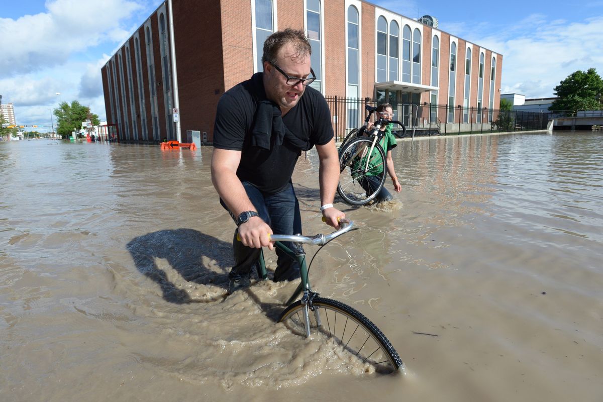 A man rides his bike as another picks his up in the flooded streets of Calgary, Alberta, on Friday. Alberta’s largest city was swamped Friday by floodwaters that submerged much of the lower bowl of the Saddledome hockey arena, displaced tens of thousands of people and forced the evacuation of the downtown core. (Associated Press)