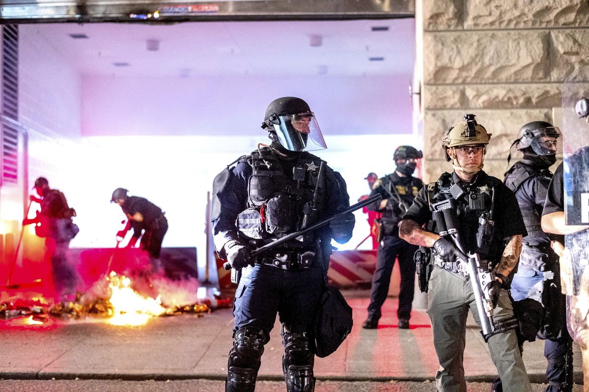 An Oregon State Police officer, right, stands watch as fellow officers extinguish a fire lit by protesters behind the Mark O. Hatfield United States Courthouse on Sun, Aug. 2, 2020, in Portland, Ore. Following an agreement between Democratic Gov. Kate Brown and the Trump administration to reduce federal officers in the city, nightly protests remained largely peaceful without major confrontations between protesters and officers.  (Noah Berger)