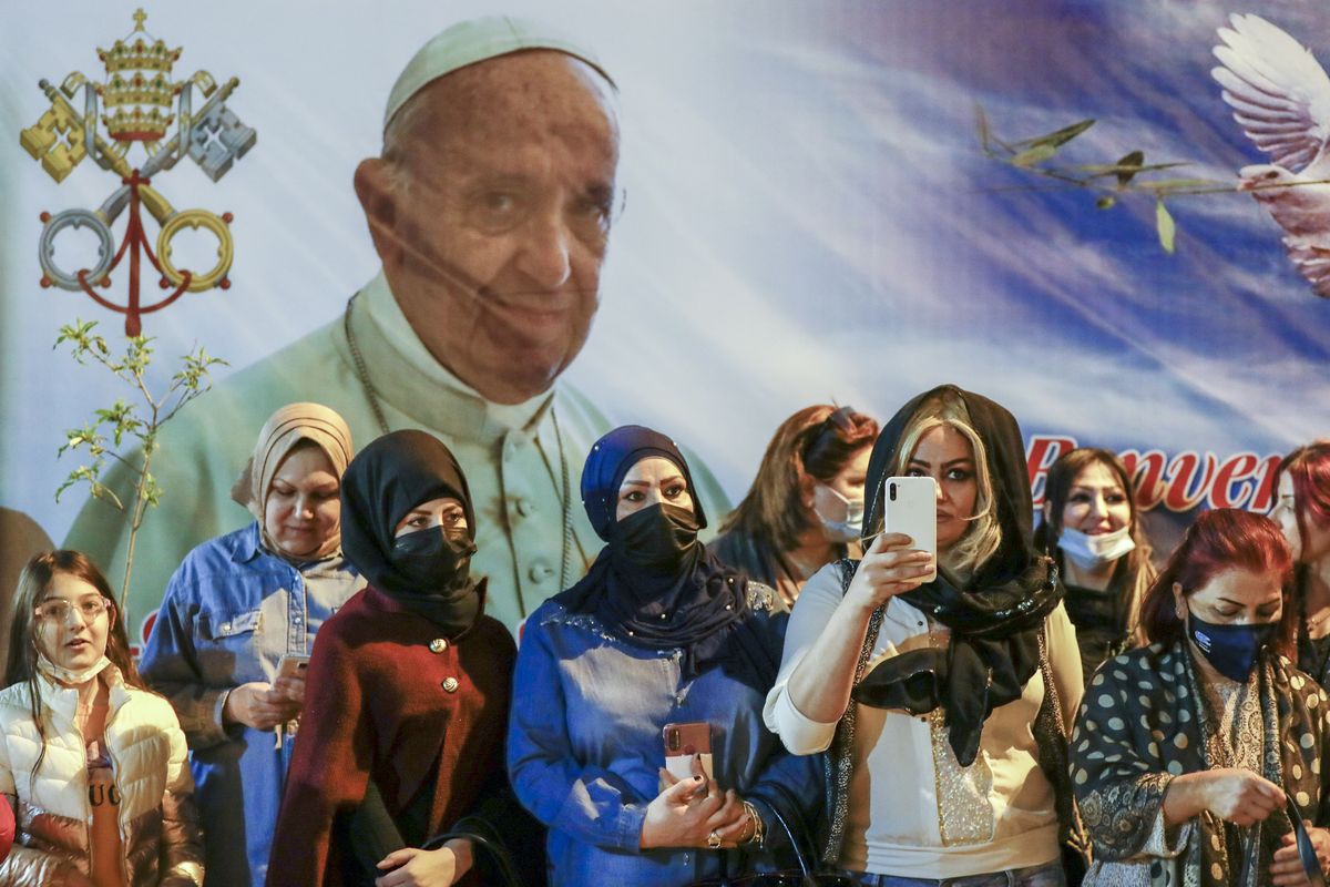 Women wait outside the Chaldean Cathedral of Saint Joseph, in Baghdad, Iraq, Saturday, March 6, 2021, where Pope Francis, depicted on a giant poster at their back, is concelebrating a mass. Earlier today Francis met privately with the country