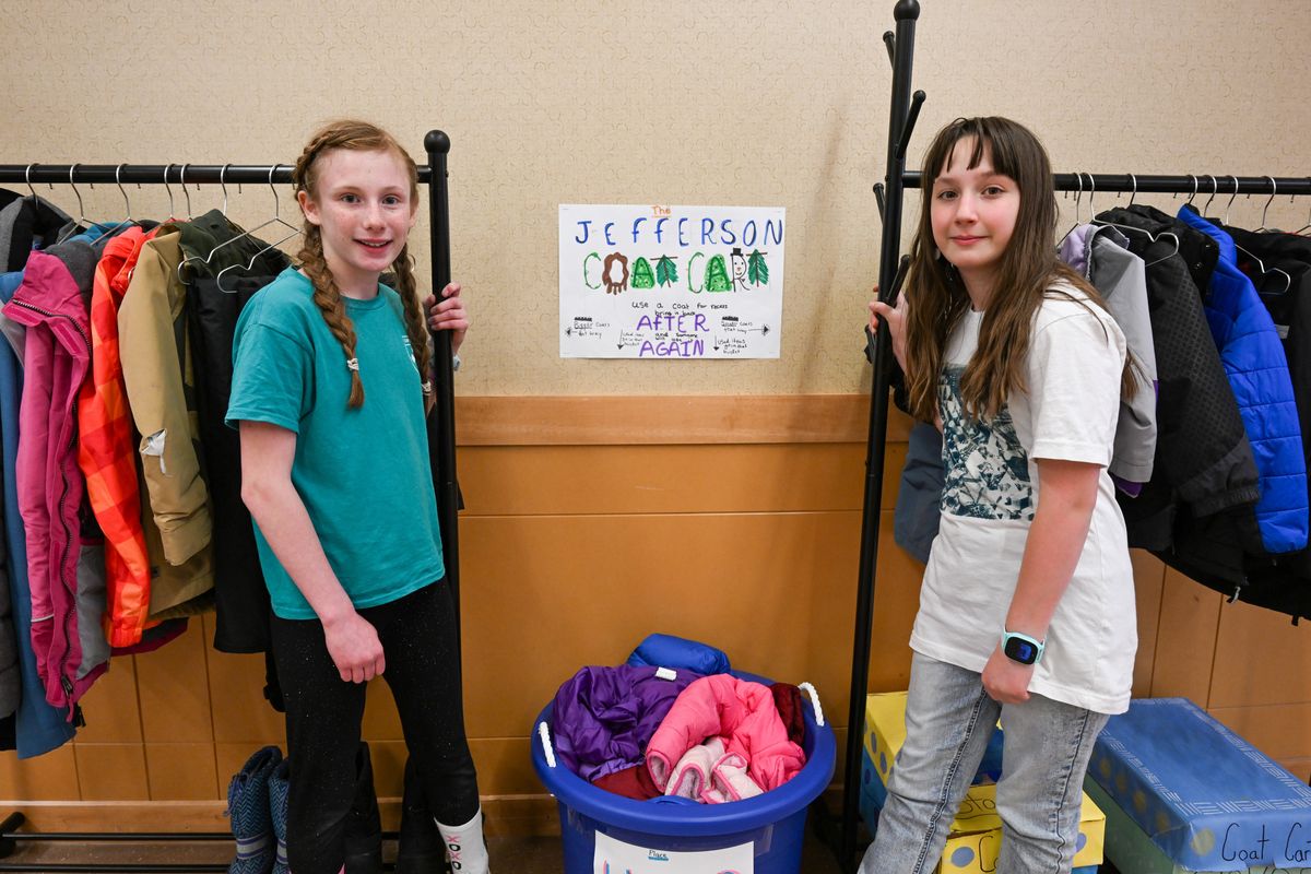 Jefferson Elementary fifth-graders Bennet Robertson, left, and Frida Sanchez stand near the Jefferson Coat Cart, an idea they came up with over the last year and is shown today in the school’s main hallway Thursday, Mar. 21, 2024 in Spokane, Washington. After watching lost and found items given away to charity, the girls came up with the idea of a coat rack carrying outdoor clothing and shoes that any kid can borrow if theyre ever not appropriately dressed for outdoor weather. The girls came up with the idea, made flyers to get donation and assembled the racks in the hallway. After they’re worn, the clothes are placed in the “used” basket to be laundered before they’re used again.  (Jesse Tinsley/THE SPOKESMAN-REVIEW)